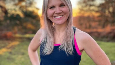 A woman standing in a garden with her hands on her hips looking into the camera wearing a gym vest