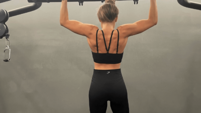 A woman facing away from the camera handing by her arms in a pull up position from gym equipment