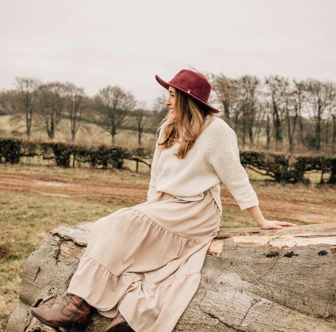 A woman facing away from the camera sitting on a felled tree wearing a hat