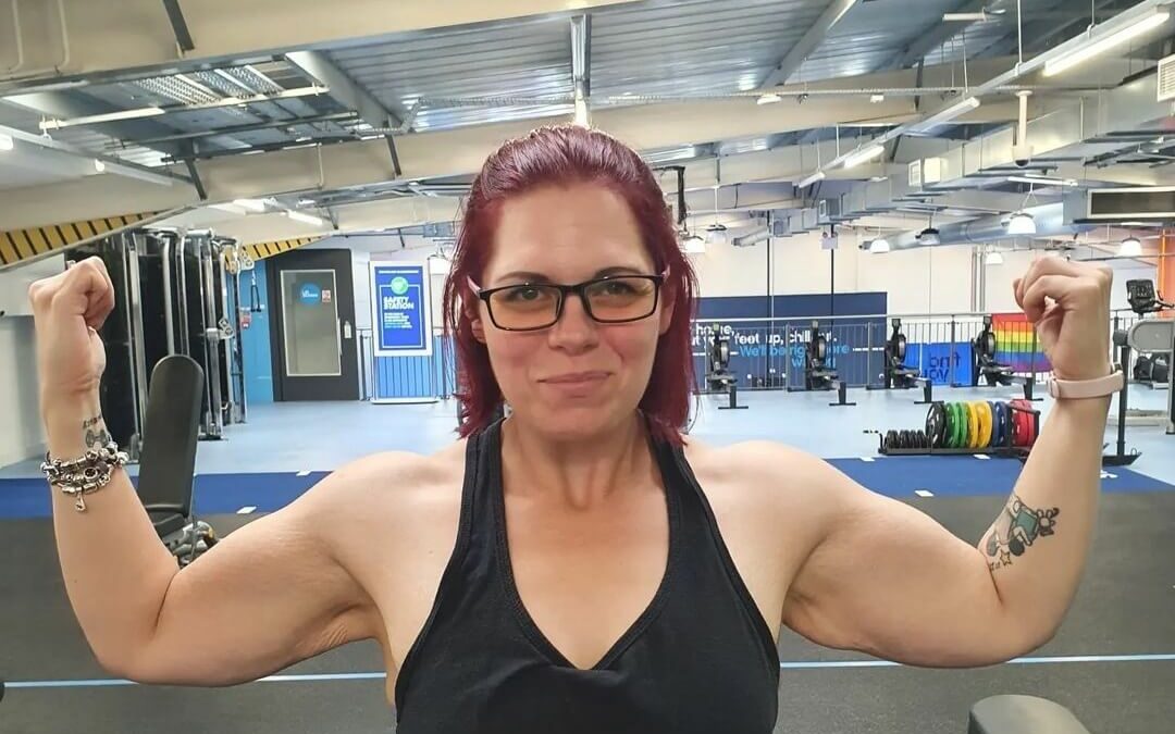 A woman in glasses and a vest flexing her arms, facing the camera