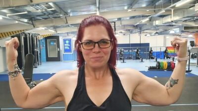 A woman in glasses and a vest flexing her arms, facing the camera